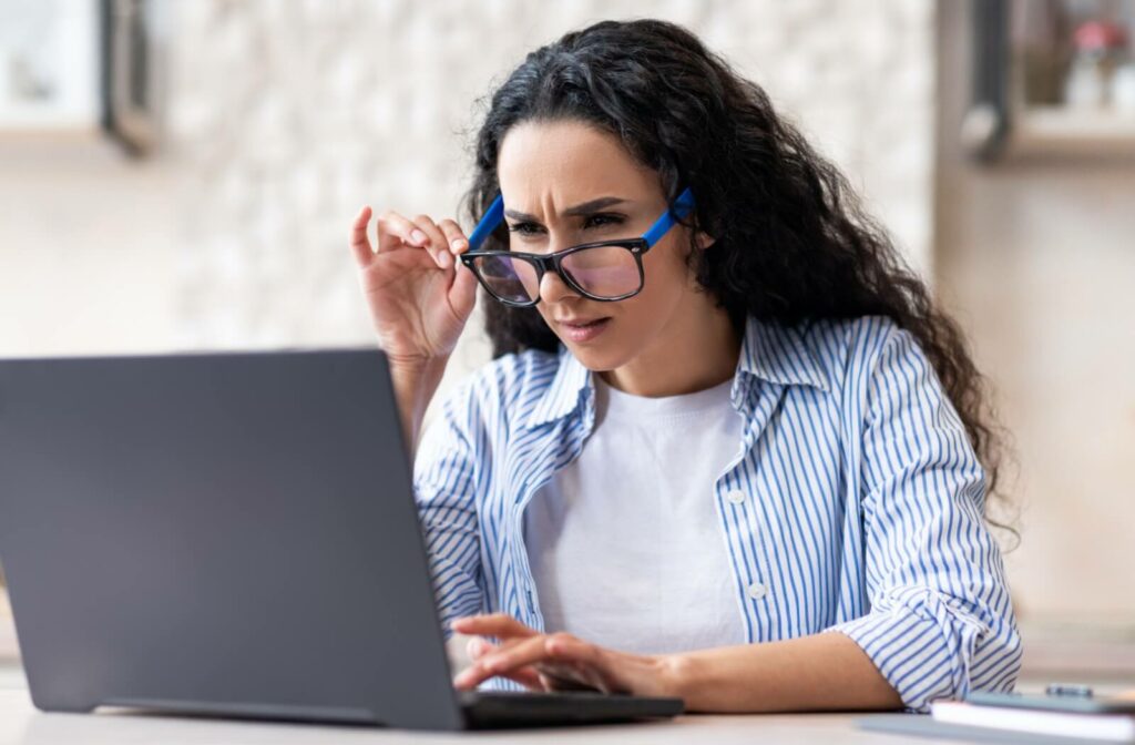 A person with blurry vision lowers their glasses and squints at their laptop screen.