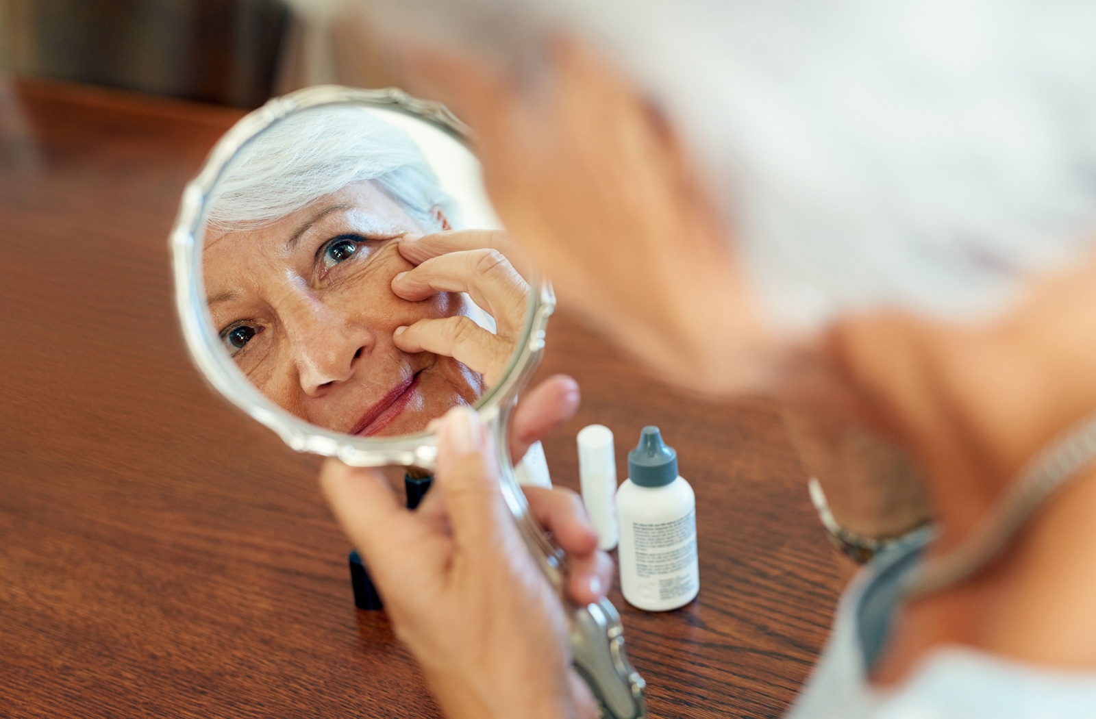 An older woman inspects her under-eye wrinkles in a hand mirror.